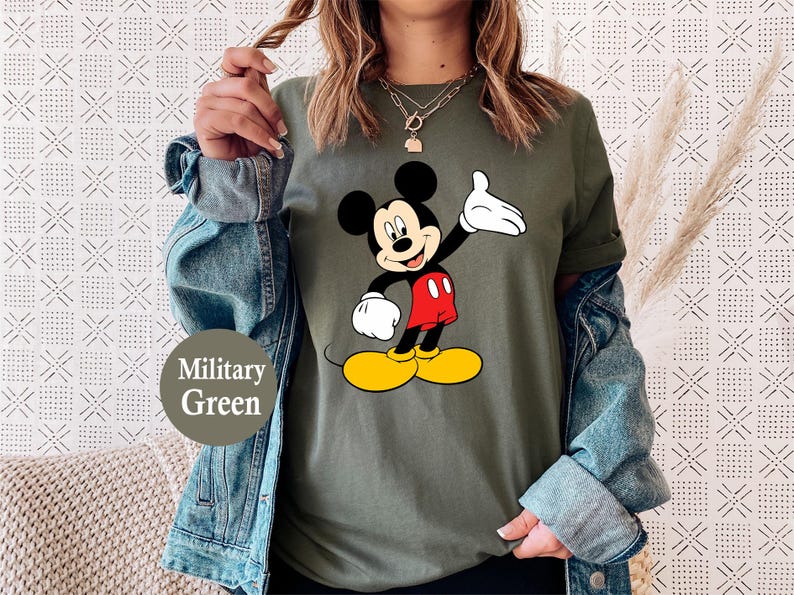 Puede incluir: Camiseta verde militar con un dise&ntilde;o de Mickey Mouse. Mickey se muestra con orejas negras, guantes blancos, pantalones cortos rojos y zapatos amarillos. El conjunto se completa con una chaqueta vaquera. Tambi&eacute;n es visible el texto "Military Green".