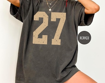 May include: Dark grey t-shirt featuring the number "27" in a tan, distressed font. The shirt has a relaxed fit and short sleeves. A black circle with the word "PEPPER" is on the right side of the shirt.
