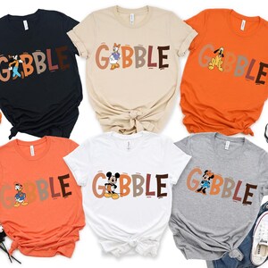 May include: A collection of six t-shirts in various colors, including black, beige, orange, white, and gray. Each shirt features the word "GOBBLE" in a playful font, with a different cartoon character incorporated into the design. The shirts are likely for Thanksgiving.
