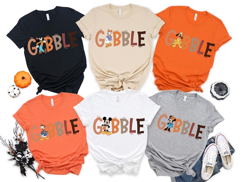 May include: A collection of six t-shirts in various colors, including black, beige, orange, white, and gray. Each shirt features the word "GOBBLE" in a playful font, with a different cartoon character incorporated into the design. The shirts are likely for Thanksgiving.