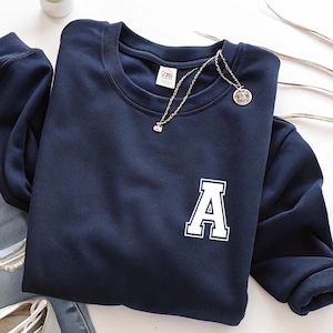 May include: Navy blue sweatshirt with a white letter "A" on the front. A silver necklace with a small pendant and a round pendant is draped across the sweatshirt. The sweatshirt is folded on a white surface.