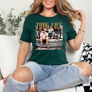 May include: A dark green t-shirt with a graphic print of a group of people sitting on a porch. The graphic includes the text "THUG LIFE" in gold letters.