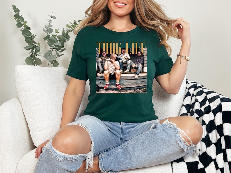 May include: A dark green t-shirt with a graphic print of a group of people sitting on a porch. The graphic includes the text "THUG LIFE" in gold letters.