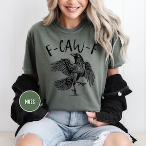 Peut inclure: T-shirt vert mousse avec un dessin de corbeau noir et le texte "F-CAW-F". Le corbeau est repr&eacute;sent&eacute; avec les ailes d&eacute;ploy&eacute;es et une patte lev&eacute;e. Le t-shirt est port&eacute; avec un jean bleu et une veste noire.