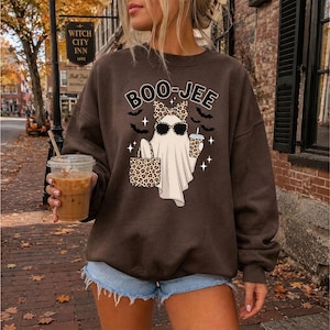 May include: A dark brown crewneck sweatshirt featuring a graphic of a white ghost wearing leopard print sunglasses and a matching bow. The ghost holds a leopard print tote bag and a drink, with the text BOO-JEE and small black bats printed above.