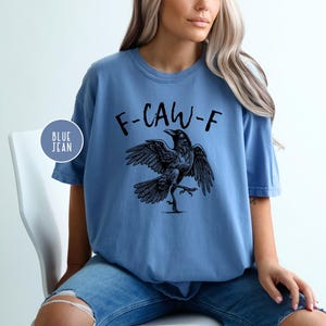 Peut inclure: T-shirt bleu clair avec l'inscription "F-CAW-F" surmontant une illustration d'un corbeau noir. Le corbeau est repr&eacute;sent&eacute; avec les ailes d&eacute;ploy&eacute;es et une patte lev&eacute;e. Le t-shirt est port&eacute; avec un jean bleu.