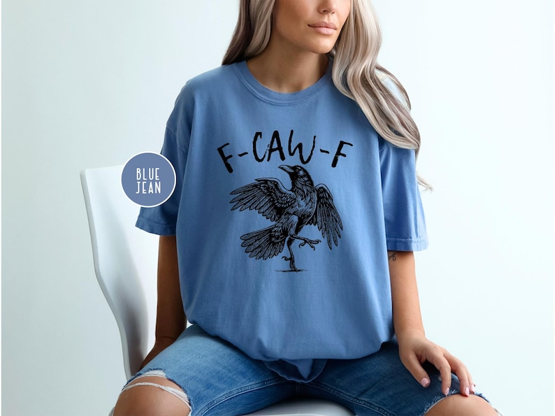 Peut inclure: T-shirt bleu clair avec l'inscription "F-CAW-F" surmontant une illustration d'un corbeau noir. Le corbeau est repr&eacute;sent&eacute; avec les ailes d&eacute;ploy&eacute;es et une patte lev&eacute;e. Le t-shirt est port&eacute; avec un jean bleu.