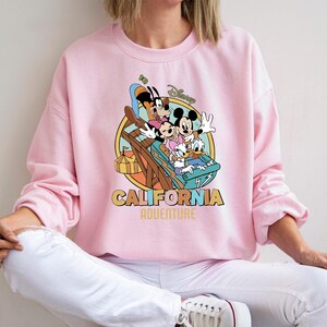 May include: Pink crewneck sweatshirt featuring a colorful graphic of Mickey Mouse, Minnie Mouse, Goofy, Donald Duck, and Daisy Duck on a roller coaster. The words "CALIFORNIA ADVENTURE" are printed below the graphic.