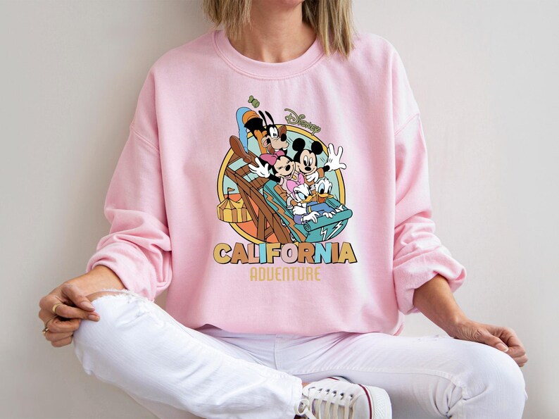 May include: Pink crewneck sweatshirt featuring a colorful graphic of Mickey Mouse, Minnie Mouse, Goofy, Donald Duck, and Daisy Duck on a roller coaster. The words "CALIFORNIA ADVENTURE" are printed below the graphic.