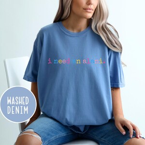 May include: A light blue t-shirt with the text "i need an alani" in rainbow colors. The shirt is made of a soft material and has short sleeves. The words "washed denim" are in a blue circle.