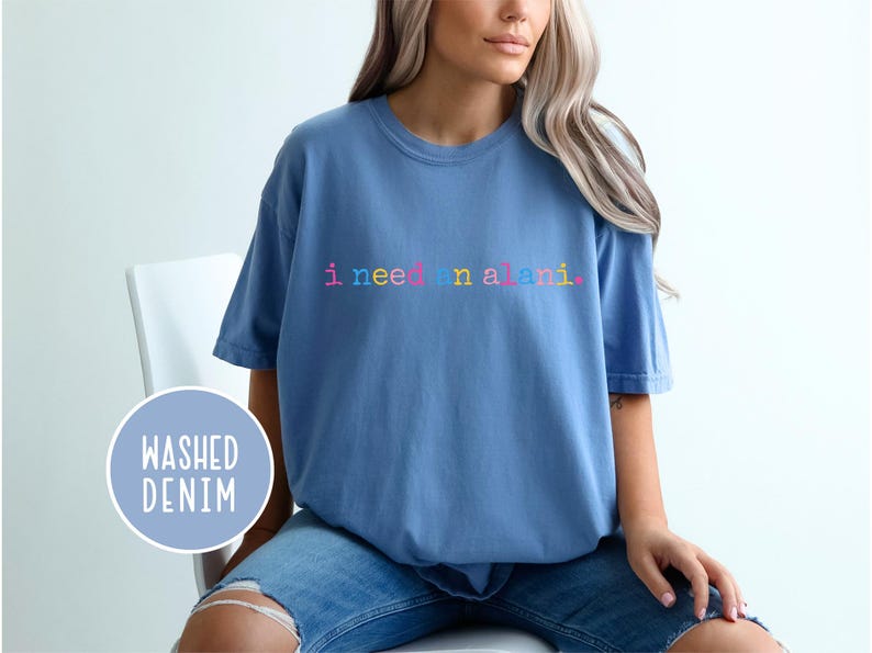 May include: A light blue t-shirt with the text "i need an alani" in rainbow colors. The shirt is made of a soft material and has short sleeves. The words "washed denim" are in a blue circle.