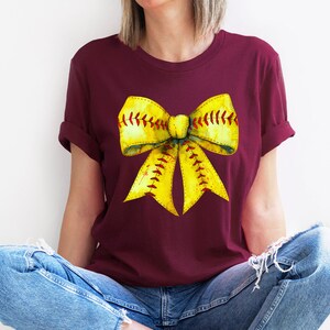 K&ouml;nnte beinhalten: Ein maroonfarbenes T-Shirt mit einem gelben Softball-Schleifendesign. Die Schleife ist so gestaltet, dass sie einem Softball mit roten N&auml;hten &auml;hnelt.