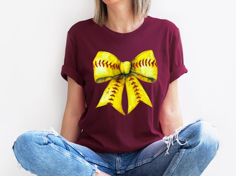 K&ouml;nnte beinhalten: Ein maroonfarbenes T-Shirt mit einem gelben Softball-Schleifendesign. Die Schleife ist so gestaltet, dass sie einem Softball mit roten N&auml;hten &auml;hnelt.