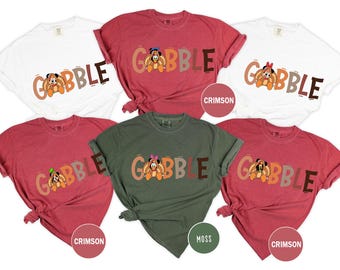 May include: A collection of t-shirts in various colors, including white, crimson, and moss green. Each shirt features the word "GOBBLE" in a colorful font, with a cartoon turkey character incorporated into the design. The shirts are likely for Thanksgiving.