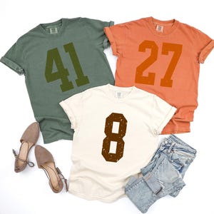 May include: Three t-shirts in olive green, orange, and cream, each with a large number printed on the front. The olive green shirt displays "41", the orange shirt displays "27", and the cream shirt displays "8". The shirts are paired with shoes and denim shorts.