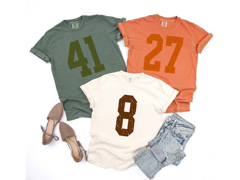 May include: Three t-shirts in olive green, orange, and cream, each with a large number printed on the front. The olive green shirt displays "41", the orange shirt displays "27", and the cream shirt displays "8". The shirts are paired with shoes and denim shorts.