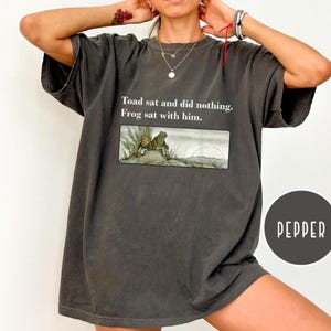 May include: Dark grey t-shirt with the text "Toad sat and did nothing. Frog sat with him." and an illustration of a toad and frog. The shirt has short sleeves and a relaxed fit. A round "Pepper" logo is visible.