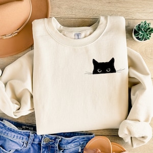May include: Cream-colored sweatshirt featuring a black cat graphic. The cat's head and eyes peek over the edge of the sweatshirt. The sweatshirt is made of a soft material and has a crew neck. The image also includes a hat, jeans, and a small plant.