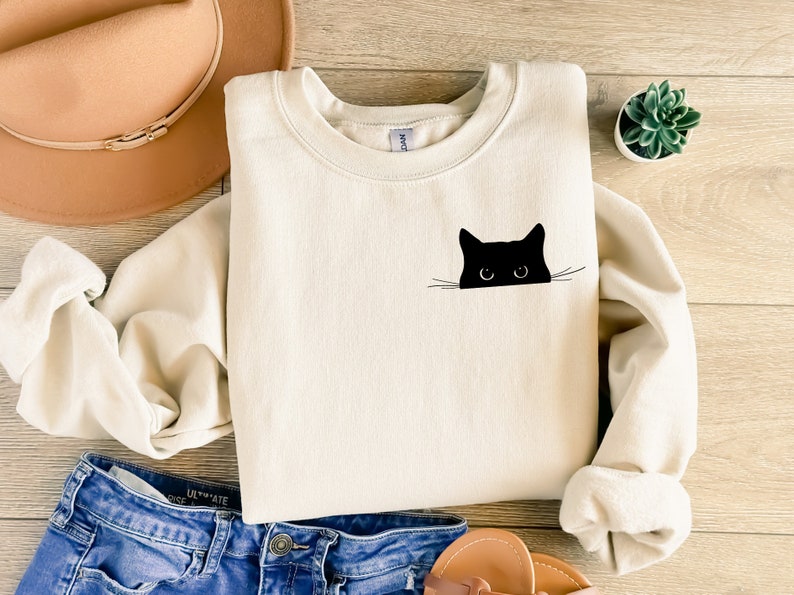 May include: Cream-colored sweatshirt featuring a black cat graphic. The cat's head and eyes peek over the edge of the sweatshirt. The sweatshirt is made of a soft material and has a crew neck. The image also includes a hat, jeans, and a small plant.