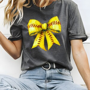 K&ouml;nnte beinhalten: Ein graues T-Shirt mit einem gelben Softball-Schleifendesign. Die Schleife ist so gestaltet, dass sie einem Softball mit roten N&auml;hten &auml;hnelt.