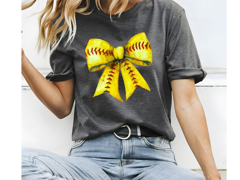 K&ouml;nnte beinhalten: Ein graues T-Shirt mit einem gelben Softball-Schleifendesign. Die Schleife ist so gestaltet, dass sie einem Softball mit roten N&auml;hten &auml;hnelt.