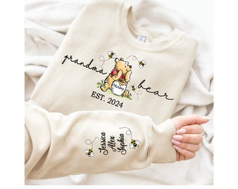 Może przedstawiać: Beżowa bluza z napisem "grandma bear EST. 2024" i kreskówkowym Winniem the Poohem trzymającym słoik miodu. Rękaw ma napis "Jessica Alex Sophia" i pszczołę.