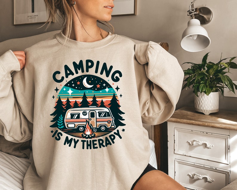 Op de afbeelding: Een beige sweatshirt met een retro-stijl grafische afbeelding van een camper in een bos met de tekst "Camping is my therapy".