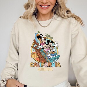 May include: Cream-colored sweatshirt featuring a colorful graphic of Disney characters Mickey Mouse, Minnie Mouse, Goofy, and Donald Duck on a roller coaster. The words "CALIFORNIA ADVENTURE" are printed below the graphic.