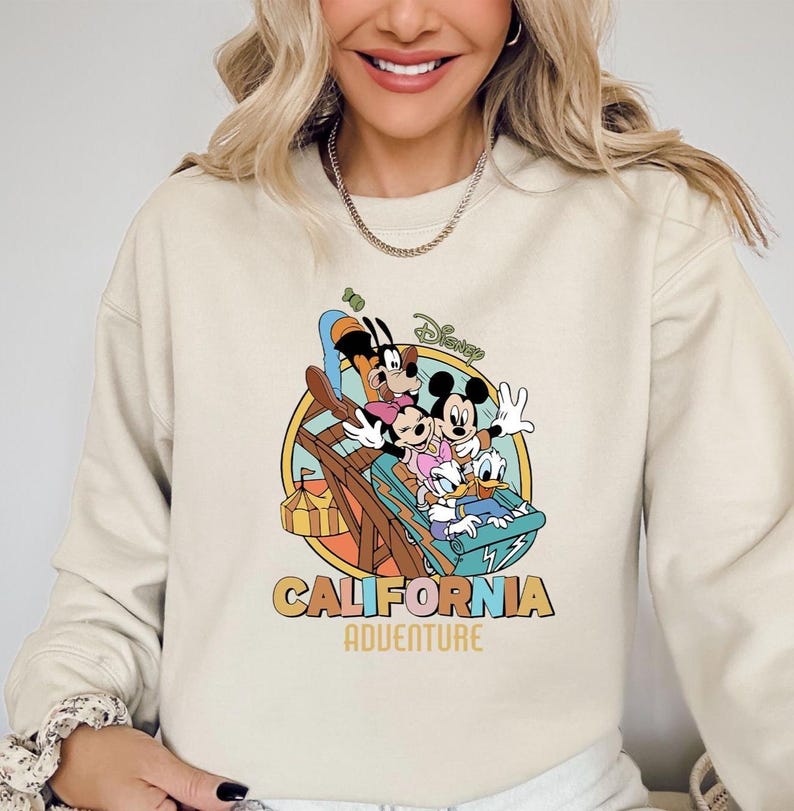 May include: Cream-colored sweatshirt featuring a colorful graphic of Disney characters Mickey Mouse, Minnie Mouse, Goofy, and Donald Duck on a roller coaster. The words "CALIFORNIA ADVENTURE" are printed below the graphic.
