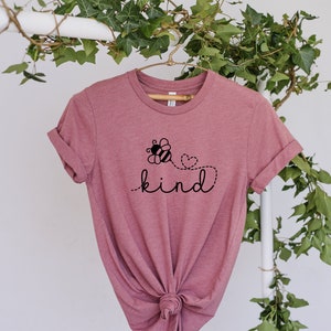 May include: A pink t-shirt with a black bee graphic and the word "kind" printed on it. The bee is flying with a heart behind it.