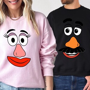May include: Pink and black sweatshirts featuring cartoon faces. The pink sweatshirt has a female face with eyelashes, large eyes, a pink nose, and a red mouth. The black sweatshirt has a male face with a mustache, orange nose, and a red mouth.