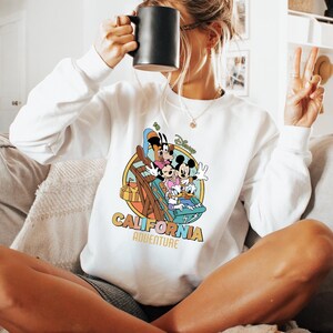 May include: White sweatshirt featuring a colorful graphic of Disney characters on a roller coaster, with the words "California Adventure" in a retro font. The sweatshirt has long sleeves and a crew neck.