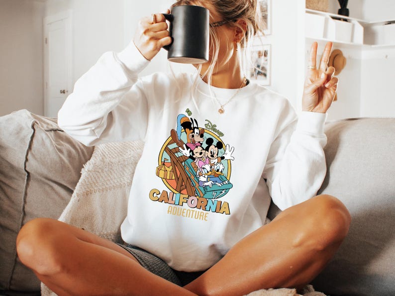 May include: White sweatshirt featuring a colorful graphic of Disney characters on a roller coaster, with the words "California Adventure" in a retro font. The sweatshirt has long sleeves and a crew neck.