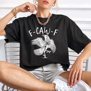Peut inclure: T-shirt noir avec un dessin blanc d'un corbeau et le texte "F-CAW-F". Le corbeau est repr&eacute;sent&eacute; avec les ailes d&eacute;ploy&eacute;es et une patte lev&eacute;e. Le t-shirt est de style d&eacute;contract&eacute; et surdimensionn&eacute;.