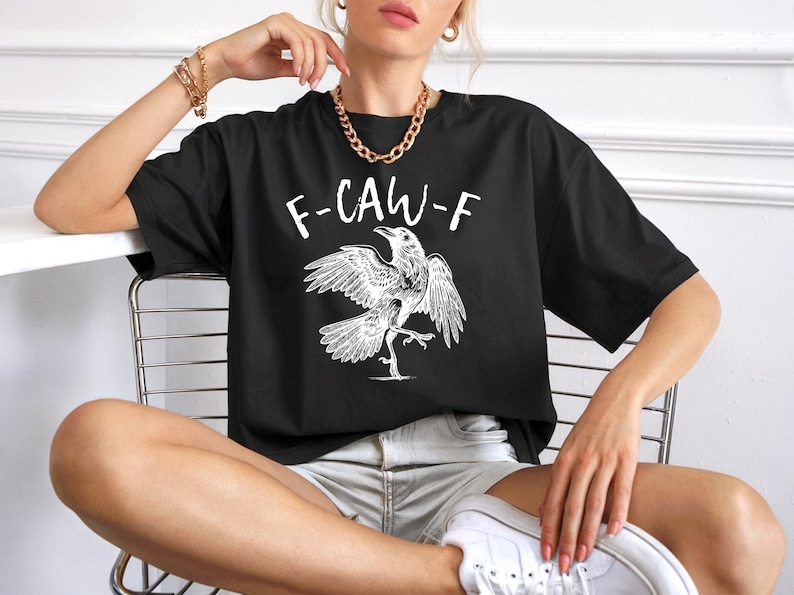Peut inclure: T-shirt noir avec un dessin blanc d'un corbeau et le texte "F-CAW-F". Le corbeau est repr&eacute;sent&eacute; avec les ailes d&eacute;ploy&eacute;es et une patte lev&eacute;e. Le t-shirt est de style d&eacute;contract&eacute; et surdimensionn&eacute;.