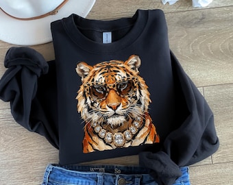 Könnte beinhalten: Ein schwarzes Langarm-Sweatshirt mit einem Grafikdruck eines Tigers, der eine Brille mit Leopardenmuster und eine große, klobige goldfarbene Halskette mit klaren, facettierten Steinen trägt, dargestellt in Orange-, Schwarz- und Weißtönen.