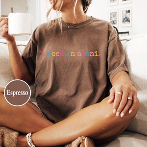 May include: Espresso-colored t-shirt with the text "i need a mani" in colorful letters. The shirt is worn by a person sitting on a couch, holding a white mug. The shirt is a casual style.