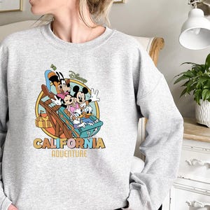 May include: A light gray sweatshirt featuring a colorful graphic of Disney characters on a roller coaster. The design includes Mickey Mouse, Minnie Mouse, Donald Duck, and Goofy, with the words "California Adventure" in a retro font.