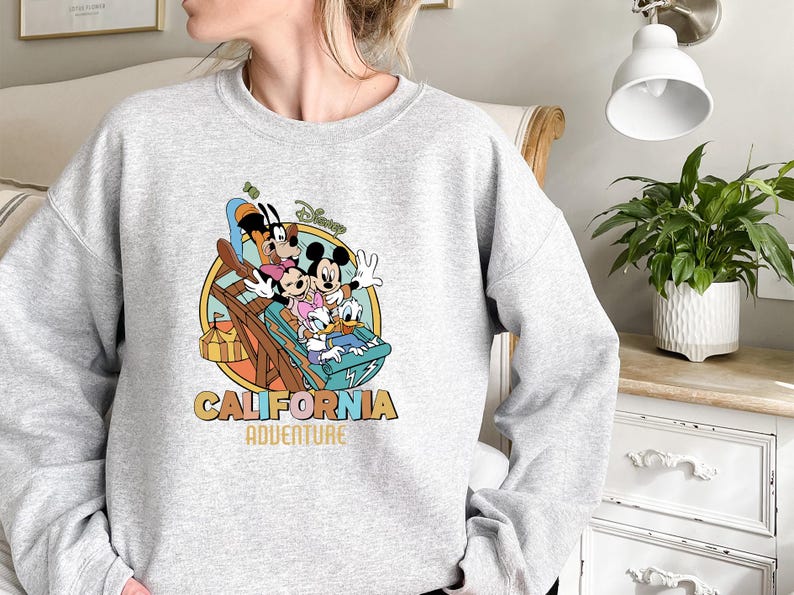 May include: A light gray sweatshirt featuring a colorful graphic of Disney characters on a roller coaster. The design includes Mickey Mouse, Minnie Mouse, Donald Duck, and Goofy, with the words "California Adventure" in a retro font.