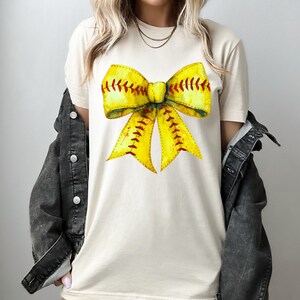 K&ouml;nnte beinhalten: Ein wei&szlig;es T-Shirt mit einem gelben und roten Softball-Schleifendesign. Die Schleife ist so gestaltet, dass sie einer Softball mit roten N&auml;hten &auml;hnelt.