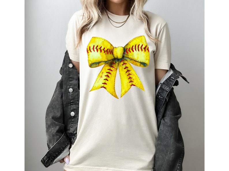 K&ouml;nnte beinhalten: Ein wei&szlig;es T-Shirt mit einem gelben und roten Softball-Schleifendesign. Die Schleife ist so gestaltet, dass sie einer Softball mit roten N&auml;hten &auml;hnelt.