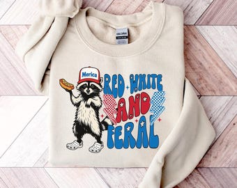 Könnte beinhalten: Cremefarbenes Sweatshirt mit einer Grafik eines Waschbären, der einen Hut mit der Aufschrift "Merica" trägt und einen Hotdog hält. Der Text "Red White and Feral" ist in Rot, Weiß und Blau gehalten. Der Waschbär trägt Crocs.