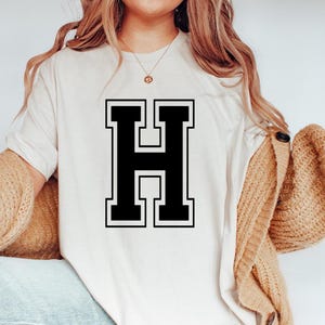May include: Cream-colored t-shirt featuring a large, black letter "H" with a white outline. The letter is centered on the shirt. The shirt is a casual style.