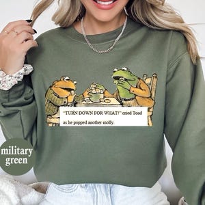 May include: Military green sweatshirt featuring a cartoon illustration of two frogs at a table. The text reads "TURN DOWN FOR WHAT!" cried Toad as he popped another molly. The sweatshirt is a solid color.