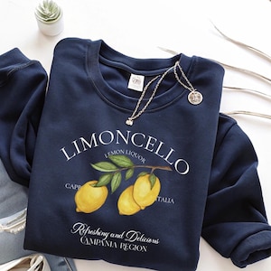 May include: A navy blue sweatshirt with the word "LIMONCELLO" in white, and a lemon branch illustration. The text "Refreshing and Delicious CAMPANIA REGION" is also displayed. A silver necklace with a pendant is draped on the sweatshirt.