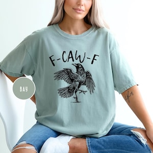 Peut inclure: T-shirt vert clair avec l'inscription "F-CAW-F" et une illustration noire d'un corbeau aux ailes d&eacute;ploy&eacute;es. Le t-shirt est ample et &agrave; manches courtes.