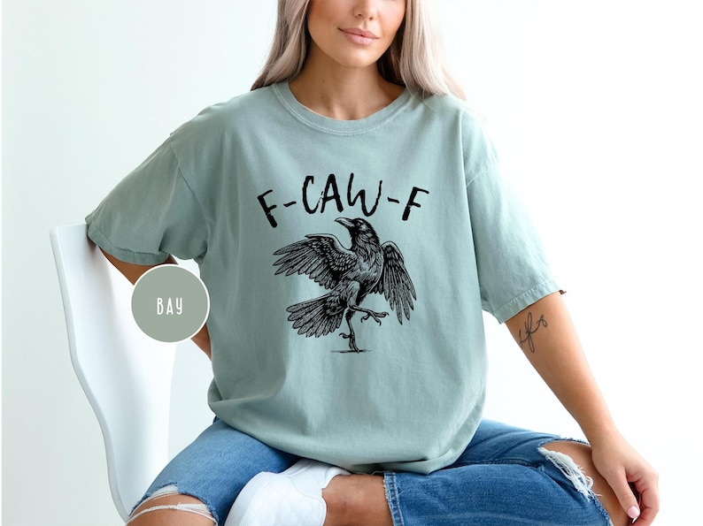 Peut inclure: T-shirt vert clair avec l'inscription "F-CAW-F" et une illustration noire d'un corbeau aux ailes d&eacute;ploy&eacute;es. Le t-shirt est ample et &agrave; manches courtes.