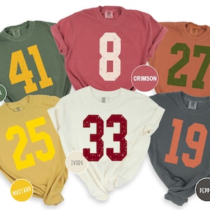 May include: Six short-sleeved t-shirts in different colours, each with a large, distressed number. Colours include green, red, orange, yellow, ivory, and grey. The numbers are in contrasting colours, such as yellow, white, and orange.