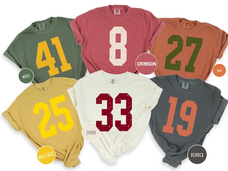 May include: Six short-sleeved t-shirts in different colours, each with a large, distressed number. Colours include green, red, orange, yellow, ivory, and grey. The numbers are in contrasting colours, such as yellow, white, and orange.