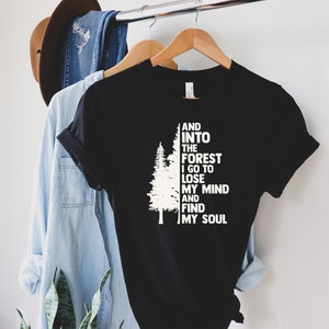 May include: Black t-shirt with white text and a tree graphic. The text reads "And into the forest I go to lose my mind and find my soul." A light blue shirt and a brown hat are visible in the background.